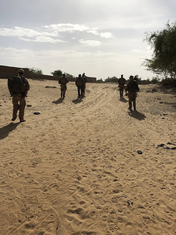Mehrere uniformierte und bewaffnete Personen gehen auf einem sandigen Weg in einer trockenen, ländlichen Umgebung. Im Hintergrund sind niedrige Gebäude und einzelne Bäume zu sehen. Die Sonne steht tief und wirft lange Schatten.